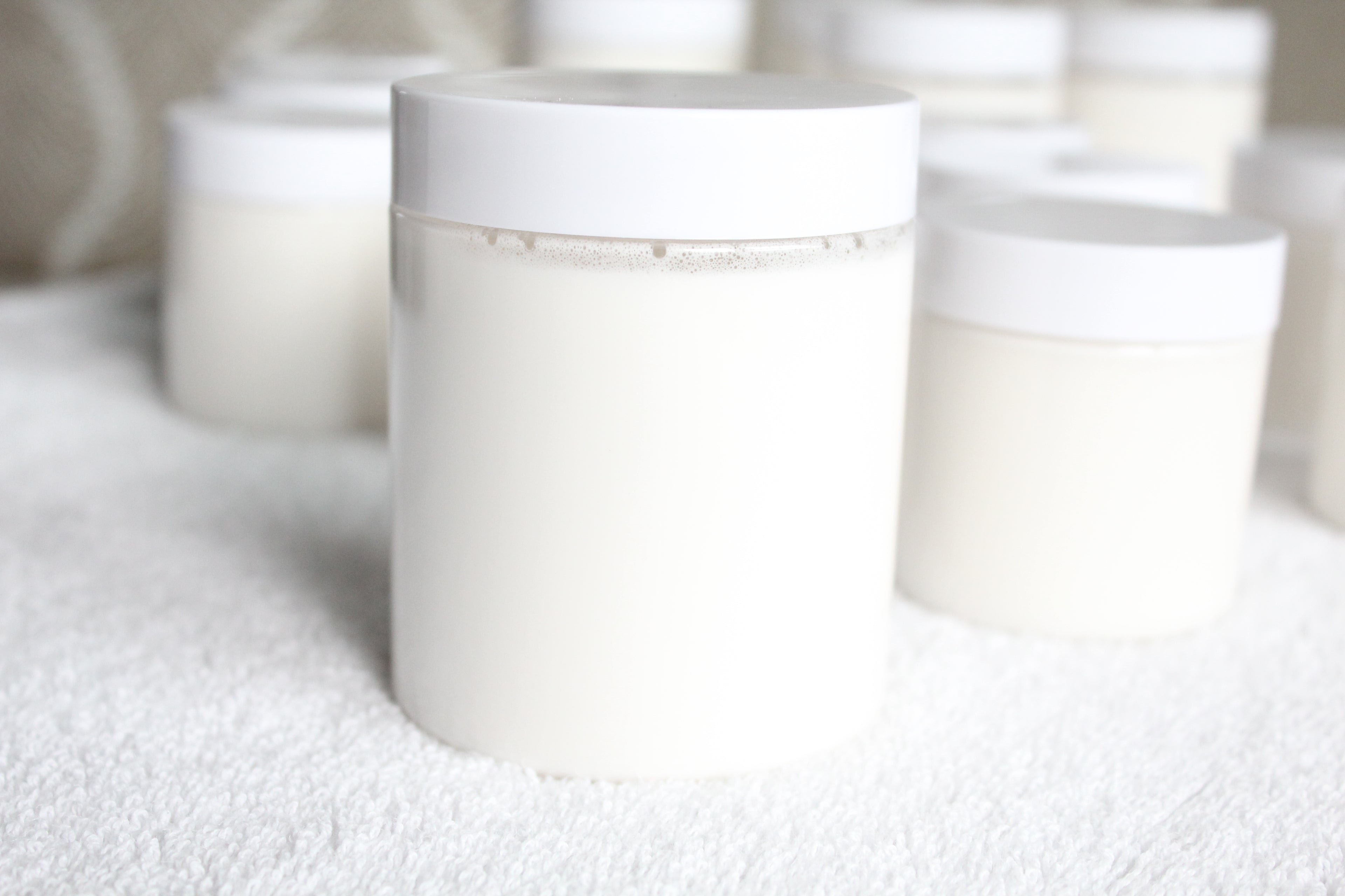 Clear plastic jar of white body butter by Busy Little Body on a white table with soft blurred background.