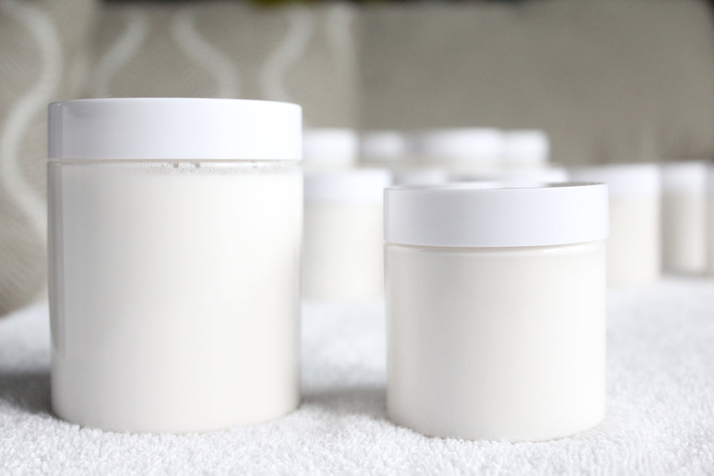 Two white plastic containers of body butter by Busy Little Body on a textured surface with a blurred background