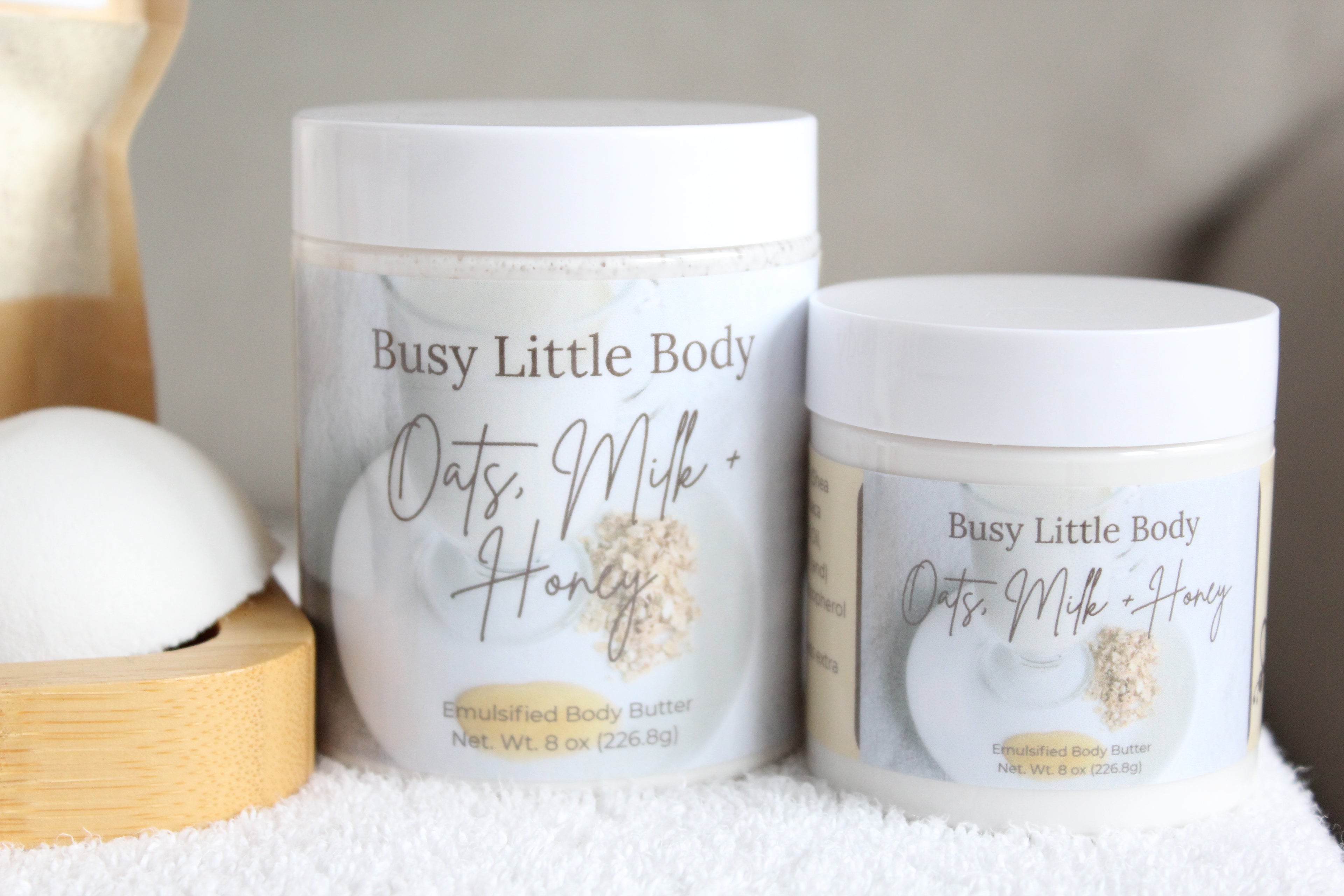 Two containers of 'Busy Little Body' Oats, Milk + Honey emollient body butter on a white surface.