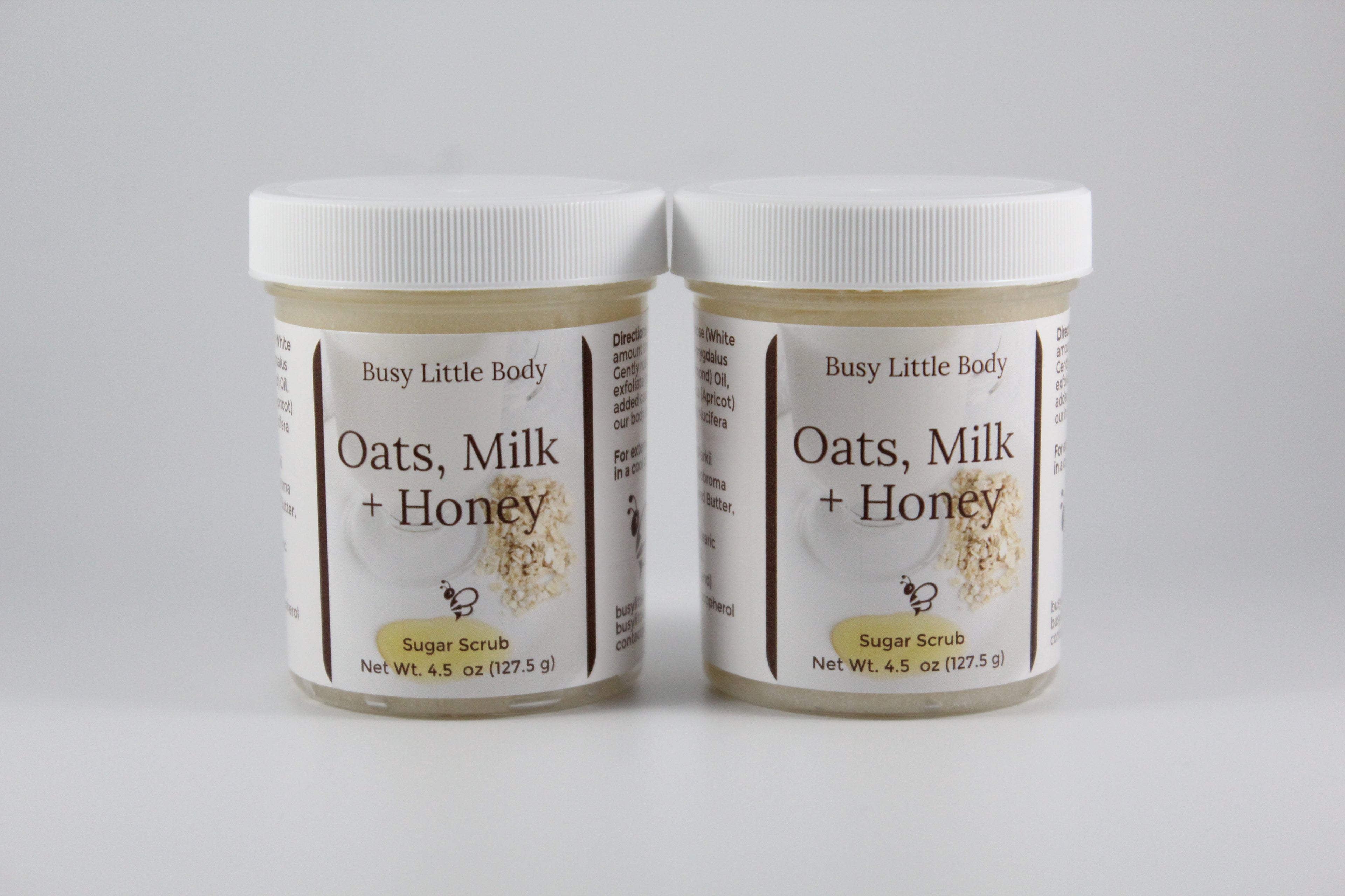 Two jars of 'Oats, Milk + Honey' sugar scrub by Busy Little Body on a gray background.