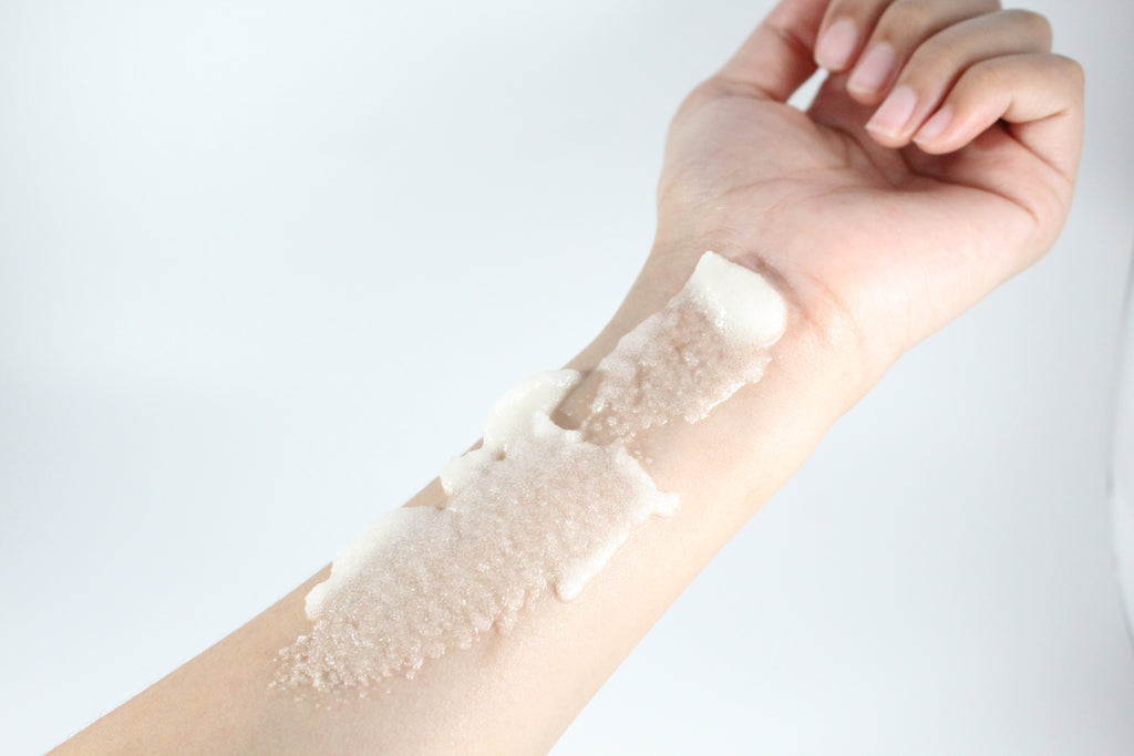 Hand applying a white sugar scrub to an arm on a light background