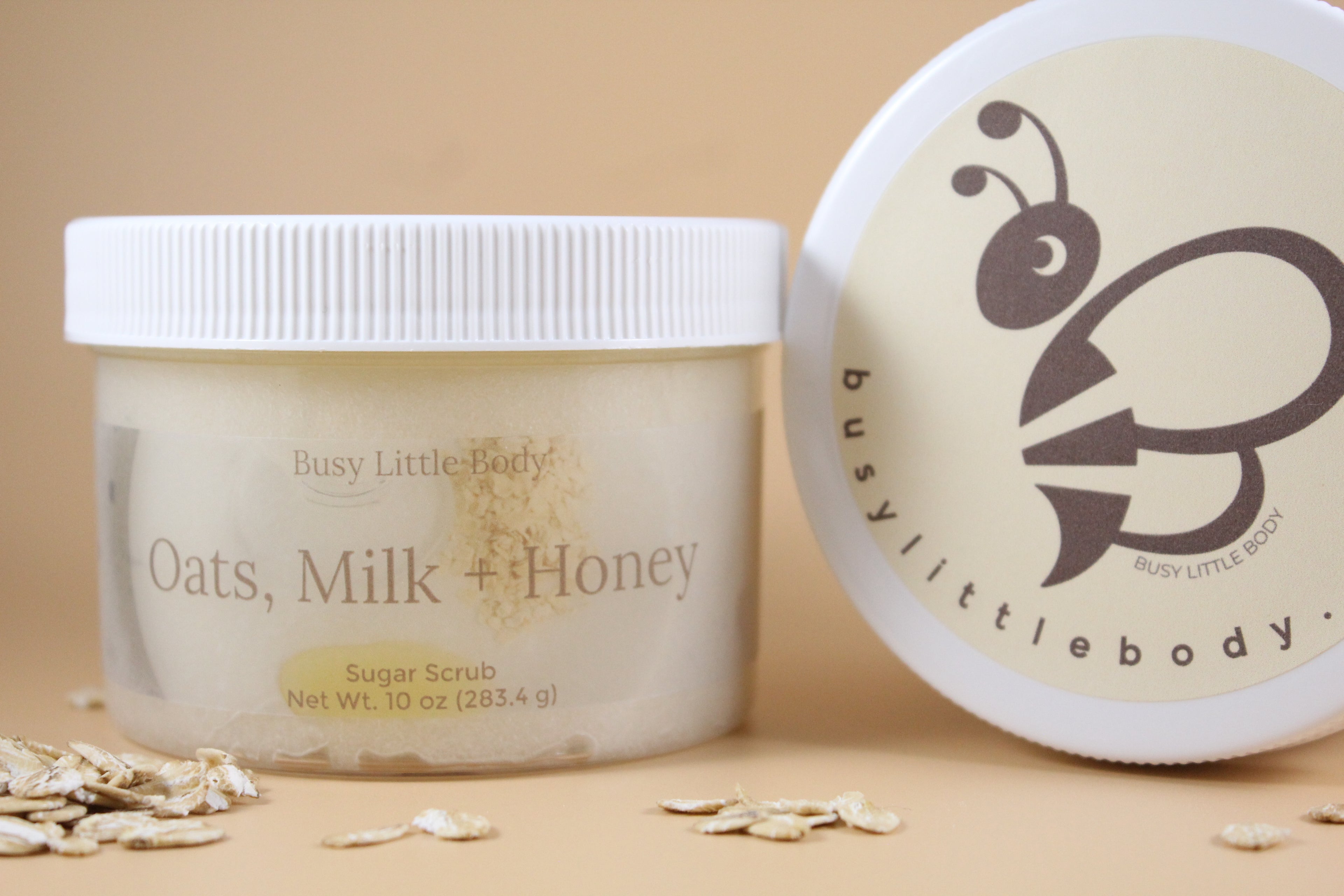 Two containers of 'Oats, Milk + Honey' sugar scrub with visible branding on a beige background.
