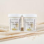 Two jars of 'Oats, Milk + Honey' body products on a soft surface with a light background.
